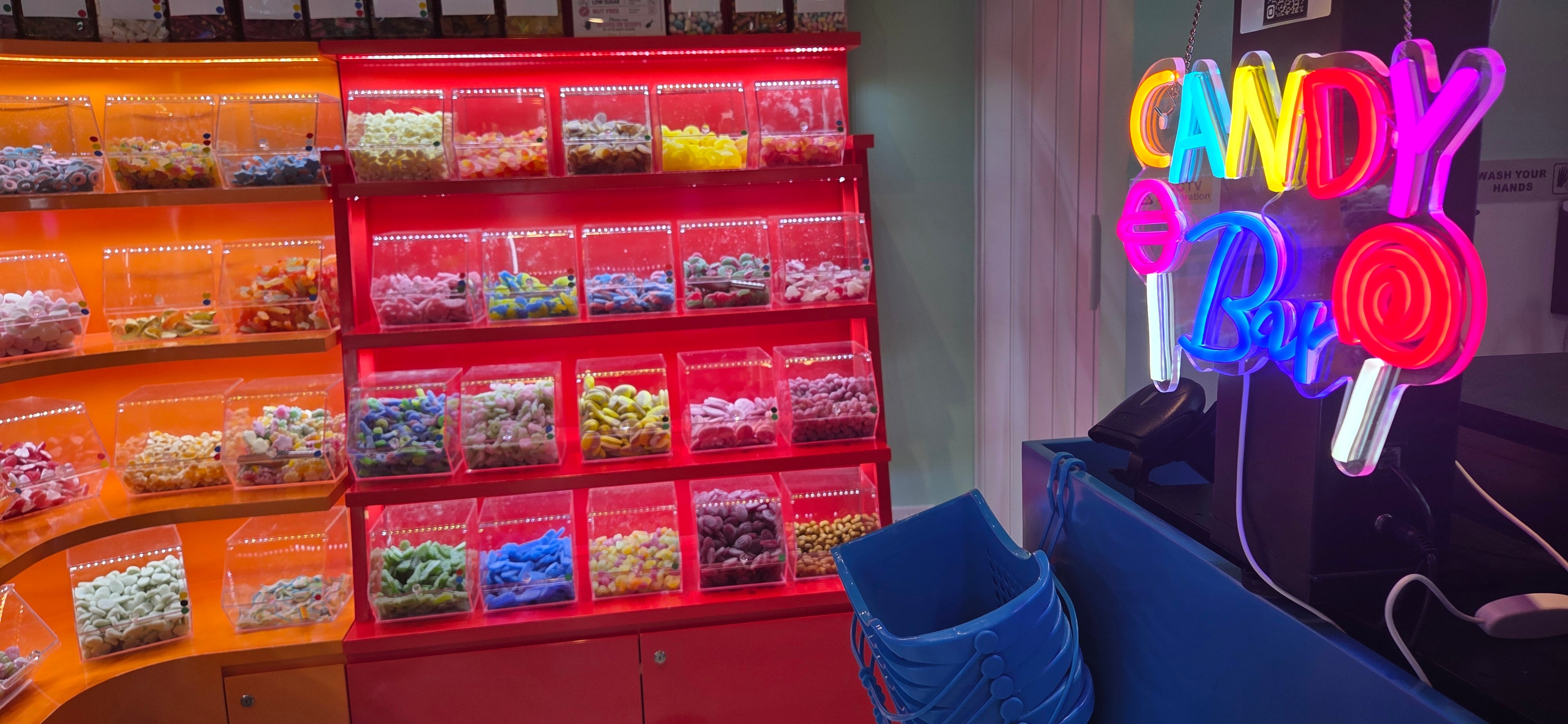  colorful candy display setup in a shop or cafe, featuring red and orange shelving filled with assorted candies, alongside a neon sign that reads 'CANDY Bar' on the right.